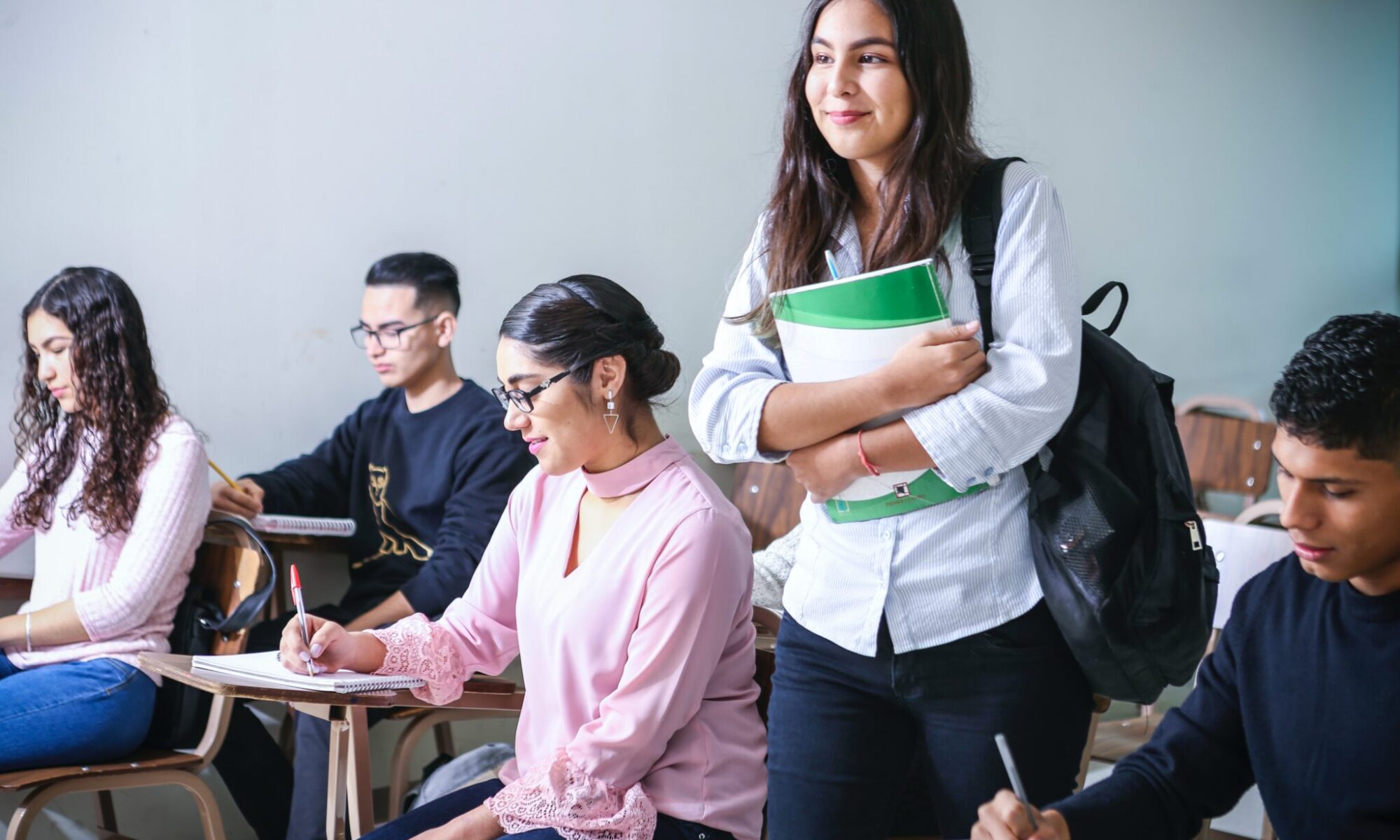 A standing college student with a backpack clutches a notebook as her peers seated nearby busily write.